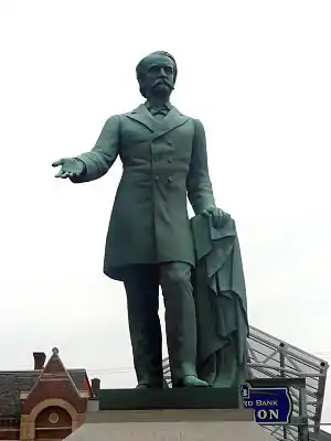A statue depicting a mustachioed man holding an overcoat in his left hand with his right hand extended