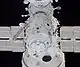 A small, cylindrical module, covered in white insulation with docking equipment at one end. In the background are some other modules and some blue solar arrays.