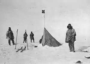  Four figures in heavy clothing stand near a pointed tent on which a small square flag is flying. The surrounding ground is ice-covered. Ski and ski poles are shown on the left.