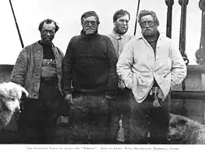  Four menwearing dishevelled clothing and worn expressions face the camera. The head of a dog is just visible on the left.